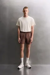 Short deportivo de entrenamiento en color marrón, confeccionado en tejido técnico ligero y elástico. Cuenta con cintura elástica ajustable con cordón, bolsillos frontales, bolsillo trasero con cierre y aberturas laterales en el bajo. Incluye un pequeño detalle de logo estampado en la pierna.