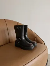 Bota de caña media estilo biker, confeccionada en cuero sintético negro. Presenta suela track robusta, hebillas metálicas laterales y detalle de argolla en el tobillo.