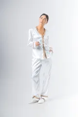 Pijama blanco de satén elastizado, compuesto por una camisa de manga larga con escote en V, abotonada adelante y con puntilla en escote y puños, y un pantalón recto con bolsillos laterales, elástico en la cintura y puntilla en el ruedo.