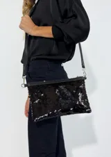 Cartera tipo sobre con lentejuelas doradas y negras reversibles, con borde superior de cuero negro y correa fina de cuero negro.