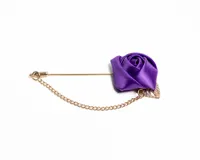 Pin para el ojal con diseño de flor de satén en color violeta, acompañada de una hoja metálica dorada y una cadena decorativa de eslabones dorados.