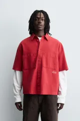 Camisa de algodón con estructura canvas, color rojo lavado, de corte relajado (relaxed fit) y manga corta. Posee cuello solapa, dos bolsillos de plastrón en el pecho y cierre frontal de botonadura. Presenta bordado y estampado de texto a contraste en el delantero.