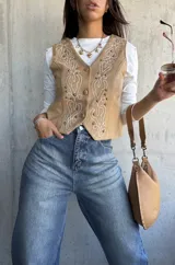 Chaleco de gamuza color beige con bordados decorativos en hilo blanco y apliques de tachas metálicas. Presenta cierre frontal con botones y diseño de corte clásico.