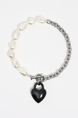 Collar asimétrico compuesto por una mitad de perlas de vidrio blancas y una mitad de cadena de eslabones plateados, con un dije de corazón negro en forma de candado y cierre de barra T.