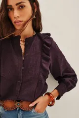 Camisa blanca de algodón con diseño romántico, cuello alto con volados, cierre frontal de botones y detalles de encaje y bordados calados. Presenta mangas largas con puños de encaje y volados frontales.