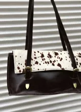 Cartera de hombro combinada en cuero liso marrón oscuro y cuero con pelo vacuno estampado tipo animal print. Presenta solapa frontal con dos correas decorativas con hebillas metálicas y detalles de herrajes en bronce.