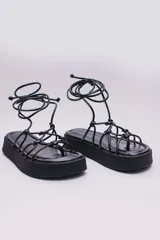Sandalias negras de ecocuero con plataforma plana y tiras finas que se atan alrededor del tobillo.