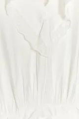 Vestido corto blanco de escote en V y manga larga con puños abotonados. Presenta volantes con bordados de encaje en el cuello, escote y falda. Cierre lateral con cremallera oculta.