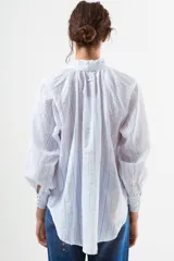Blusa de algodón blanco con diseño de rayas verticales y textura de pequeños puntos en relieve. Presenta cuello alto con volados, cierre frontal con botones forrados y mangas largas abullonadas con puños ajustados.