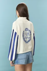 Cardigan de algodón color azul con mangas largas de rayas verticales anchas en tonos azul y celeste, puños en rojo, y bordado en la espalda con diseño de escudo nacional.