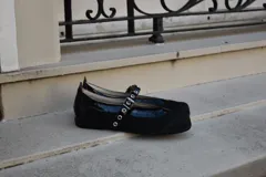 Ballerina Ines de tela negra con detalles de cuero y forrería vacuna. Presenta tira con hebilla ajustable y ojalillos metálicos sobre el empeine.