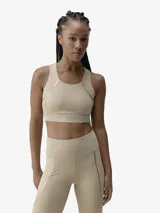 Top deportivo de color beige con diseño de escote redondo y tirantes anchos. Presenta detalles de cintas doradas termoselladas en los laterales y una banda elástica inferior con logo sutil. Confeccionado en tejido técnico compacto y ligero con copas extraíbles.