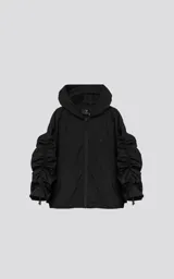 Campera rompevientos negra de calce oversize, confeccionada en microfibra técnica ligera con acabado semi-mate. Presenta capucha integrada, cierre frontal técnico y bolsillos tipo ojal. Destaca por su sistema de frunces elásticos regulables mediante tancas a lo largo de las mangas y el ruedo, permitiendo ajustar el volumen de la prenda.