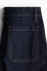 Jean de tiro alto en denim azul oscuro, con corte acampanado desde la rodilla. Presenta un diseño con cordones frontales para ajuste en la cintura, bolsillos delanteros y traseros, y costuras a contratono.