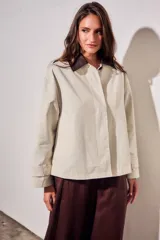 Trench corto de corte oversize con estampado de cuadros finos en tonos beige y verde. Presenta cuello camisero en contraste de color marrón oscuro, cierre frontal con botones y mangas largas con detalle de trabas en los puños.