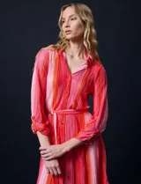 Vestido camisero midi de satén plisado con estampado a rayas verticales en tonos rosa y naranja. Tiene cuello camisero, cierre frontal con botones, mangas largas con puños abotonados y cinturón de lazo.