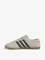 Zapatillas Adidas Gazelle Lo Pro color gris claro con las tres tiras laterales en negro y suela de goma color caramelo.