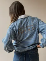 Buzo tipo chomba de corte oversize con diseño a rayas horizontales celestes y blancas. Presenta cuello camisero blanco y un bordado en la espalda con la inscripción República Oriental del Uruguay.
