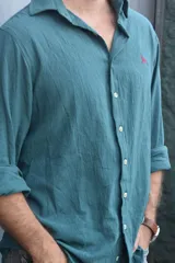 Camisa de manga larga, color verde azulado (verde inglés), con cuello camisero y botones frontales. Presenta un pequeño logo bordado en color rojo en el pecho. El tejido parece ser de lino o una mezcla ligera y arrugada.
