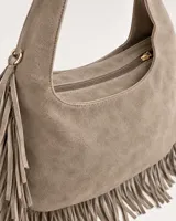 Cartera estilo hobo de gamuza color beige, con flecos largos colgantes en los laterales y la base. Cuenta con asa ancha integrada y cierre superior con cremallera.