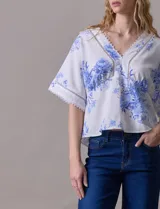 Blusa blanca estampada con flores azules, escote en V y mangas cortas amplias. Detalles de puntilla en escote y mangas.