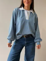 Buzo tipo chomba de corte oversize con diseño a rayas horizontales celestes y blancas. Presenta cuello camisero blanco y un bordado en la espalda con la inscripción República Oriental del Uruguay.