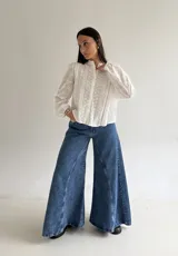 Blusa de brodery color blanco, con cuello redondo, mangas largas abullonadas con puños elásticos y cierre frontal con botones forrados. Presenta detalles de puntillas verticales en el frente.