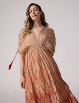 Vestido largo de gasa color salmón con un sutil patrón estampado brillante en tono dorado. Cuenta con escote en V cruzado, cintura imperio elastizada y mangas cortas con tiras ajustables terminadas en borlas.