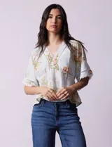 Blusa blanca estampada con flores azules, escote en V y mangas cortas amplias. Detalles de puntilla en escote y mangas.