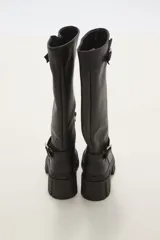 Bota de caña alta color negro, confeccionada en eco cuero, con suela track de goma y detalle de hebilla metálica en el tobillo y en la parte superior de la caña.