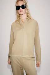 Polo oversize de algodón color beige, de manga larga, con cuello camisero y cierre frontal de botones ocultos.