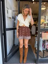 Shorts de tiro alto color marrón con estampado de pequeños rombos blancos y un borde ancho con diseño de encaje o bordado estilo paisley en blanco.