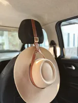 Accesorio porta sombrero de cuero con correa ajustable y hebilla metálica, diseñado para colgarse en el apoyacabezas del automóvil y transportar sombreros de forma segura.