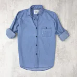 Camisa celeste de manga larga con cuello clásico, cierre con botones y bolsillo en el pecho con detalle de rotura.