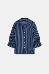 Camisa de rayas verticales azules y blancas, de cuello solapa con escote en V y cierre frontal con botones. Presenta mangas abullonadas por debajo del codo.