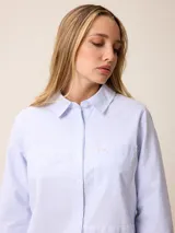Camisa de manga larga con cuello clásico y cierre frontal de botones, confeccionada en tela de algodón con diseño de rayas verticales finas en color rosado y blanco. Presenta un bolsillo frontal en el pecho con detalle de etiqueta pequeña.