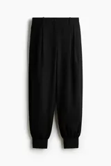 Pantalón negro de tiro medio con corte holgado y diseño de piernas globo, pinzas en la parte delantera y puños elásticos ajustados en los tobillos. Cuenta con cierre frontal y trabillas para cinturón.