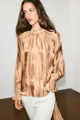 Camisa satinada color tostado con cuello alto fruncido, mangas largas amplias y puños elásticos con volados.