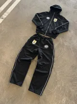 Campera deportiva negra con capucha, cierre frontal metálico y detalles de ribetes blancos. Presenta un escudo bordado en el pecho con motivos uruguayos y un logo reflectivo circular en el lado opuesto. Cuenta con bolsillos laterales con cierre.