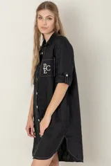 Vestido camisero negro de lino con botones, mangas 3/4 con trabilla y bolsillo de parche en el pecho con bordado de letras. Tiene aberturas laterales en el ruedo y costuras a contratono en color crudo.
