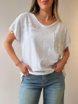 Remera blanca de lino y algodón, de corte relajado con escote en V y mangas cortas. Presenta estampado de escritura manuscrita, bolsillo aplicado con detalles de encaje, strass y tachas, y terminaciones desflecadas.