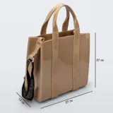 Cartera tipo tote bag Melissa modelo Dulce Bag, color tostado, fabricada en plástico PVC brillante. Cuenta con asas de mano fijas, tiras decorativas verticales con el logotipo de la marca y una correa larga ajustable y desmontable en color negro con inscripciones.