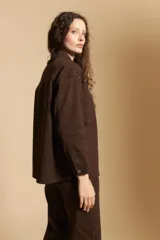 Sobrecamisa de gabardina color marrón, con cierre frontal de botones, bolsillos en el pecho y detalle de tachas metálicas en los hombros y canesú. Presenta cuello clásico y mangas largas con puños abotonados.