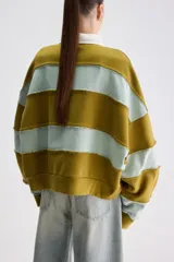 Buzo de estilo rugby con diseño oversize, confeccionado en bloques de color verde militar y celeste con costuras expuestas y deshilachadas. Cuenta con cuello camisero blanco de popelina y cartera de botones frontal oculta, además de mangas largas y puños acanalados.