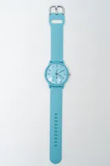Reloj analógico con correa de silicona color celeste claro y esfera del mismo tono. La esfera presenta números y marcas de hora en un tono ligeramente más claro, con tres manecillas y tres subesferas decorativas. La correa tiene un cierre de hebilla metálico y una hilera de perforaciones.
