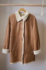 Campera tipo gamulán de cuero genuino en color marrón, con interior y detalles en corderito sintético color crema. Presenta cierre frontal con botones, cuello solapa y puños con vuelta.