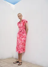 Vestido midi de cuello camisero con cierre frontal de botones, manga corta y cinturón ajustable en la cintura. Presenta un diseño recto con estampado floral en tonos fucsia y rosado.