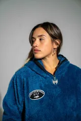 Canguro de textura tipo peluche en color azul, con cierre frontal parcial, cuello alto y bolsillo tipo canguro. Presenta un parche ovalado en el pecho con diseño gráfico.