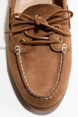 Zapatos náuticos de gamuza color marrón, con cordones frontales decorativos, costuras visibles en la puntera y ojalillos metálicos dorados. Incluyen forro interno.