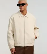 Campera masculina de corte boxy en color beige, con cuello inglés, cierre frontal de cremallera, mangas largas con puños elásticos y ruedo con elástico. Presenta bolsillos laterales y detalles de recortes en el diseño.